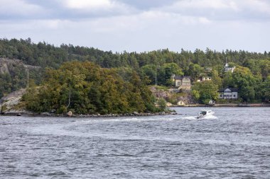 Yeşillik ve ön planda gezinen bir teknenin olduğu küçük bir adanın manzarası. Arka planda bulutlu bir gökyüzünün altında büyüleyici evleri ve ağaçları olan bir kıyı şeridi yer alıyor..