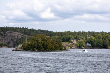 Bir adanın yanından geçen küçük bir tekneyle sakin bir su cesedinin manzarası. Ada yeşilliklerle doludur ve görünür birkaç evi vardır. Arka planda, bulutlu bir gökyüzünün altında kayalık kayalıklar ve yoğun ağaçlar var..