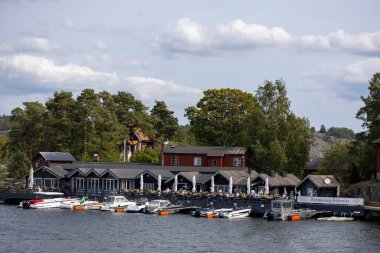 Sahile yanaşmış ağaçlar ve teknelerle çevrili açık deniz manzaralı bir restoran. Gökyüzü parçalı bulutlu, sakin bir atmosfer yaratıyor..