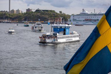 Suda dolaşan çeşitli tekne ve feribotların yer aldığı yoğun bir liman sahnesi. Ön planda, 'Djurgrden 8' adlı bir feribot belirgin bir şekilde görünür, diğer gemiler ve bir yolcu gemisi ise arka planda. 