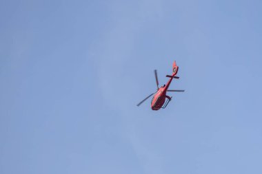 Mavi gökyüzünde uçan kırmızı bir helikopter, hareket halindeki pervanelerini gösteriyor. Görüntü helikopteri aşağıdan yakalıyor, canlı rengini gökyüzünün arka planına vurguluyor..