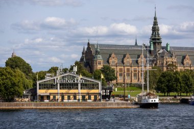 Stockholm 'de bir çocuk müzesi olan Junibacken' in manzarası. Arka planda tarihi bir bina ve önde de yemyeşil ve açık mavi gökyüzüyle çevrili bir yelkenli..