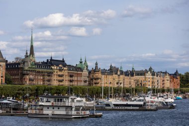 Şehrin rıhtımının manzarası. Süslü mimarisi olan tarihi binalar, ön planda demirli tekneler ve bulutlu bir gökyüzü..