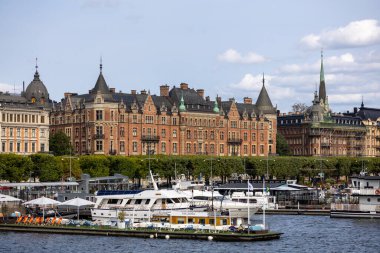 Stockholm 'de tarihi tuğla binaların, kulelerin ve kulelerin bulunduğu bir rıhtım manzarası. Tekneler ön planda, açık mavi bir gökyüzü ve kabarık bulutlarla kenetlendi..