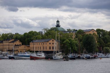 Bereketli yeşillikler ve tarihi binalarla çevrili çeşitli teknelerle dolu bir limanın manzarası. Gökyüzü bulutlu ve manzaraya dramatik bir etki yapıyor..