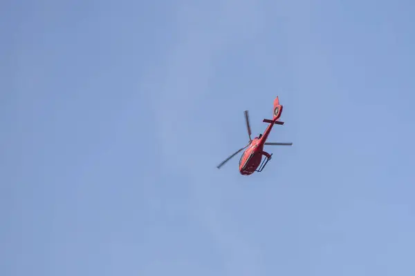 Mavi gökyüzünde uçan kırmızı bir helikopter, hareket halindeki pervanelerini gösteriyor. Görüntü helikopteri aşağıdan yakalıyor, canlı rengini gökyüzünün arka planına vurguluyor..