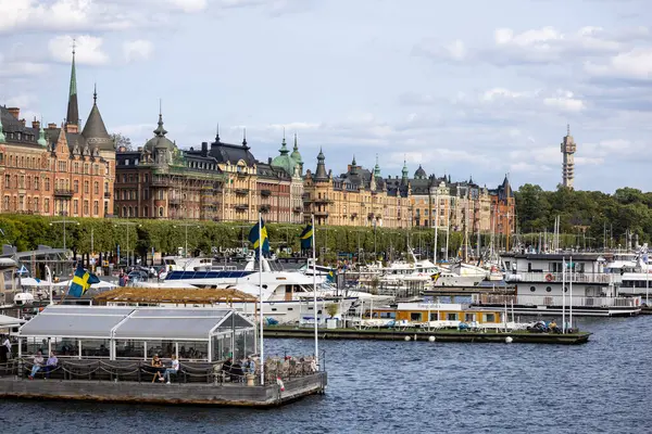 Stockholm, İsveç 'te tarihi binalar, tekneler ve İsveç bayraklarının yer aldığı bir rıhtım manzarası. Gökyüzü parçalı bulutlu, resim atmosferini güçlendiriyor..