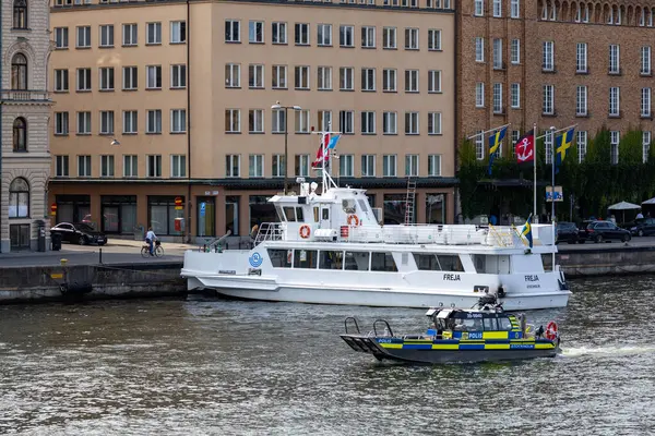 Freja adında beyaz bir yolcu teknesi nehir kıyısına yanaşmış, yanında da daha küçük bir polis botu var. Arka planda, canlı bir kent ortamını yansıtan modern binalar ve bayraklar yer alıyor..