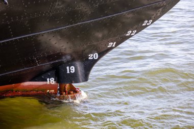 Geminin gövdesine yakın çekim. Görünür sayıda cereyan izi var. Suya batmış. Gövde siyah, paslı kırmızı bir tabanı var ve su sakin..