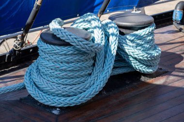 İki siyah krampon üzerinde dinlenen, bir tekne güvertesinde sarmalanmış kalın, mavi denizcilik halatı yakın plan. Arka planda mavi bir yelken ve ahşap bir güverte var..