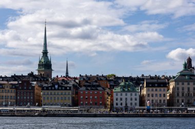Stockholm 'ün rıhtımının manzarası. Parçalı bulutlu bir gökyüzünün altında renkli tarihi binalar sergileniyor. Durgun su, canlı bir mimariyi yansıtıyor ve pitoresk bir şehir manzarası yaratıyor..