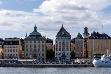 Stockholm, İsveç 'teki rıhtım boyunca uzanan renkli tarihi binaların manzarası. Önünde tekneler ve parçalı bulutlu bir gökyüzü var..