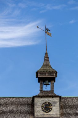 Üstünde rüzgar gülü olan bir saat kulesinin yakından görüntüsü. Zil ve saat yüzlü. Gökyüzü mavi ve arkaplanda ince bulutlar var..