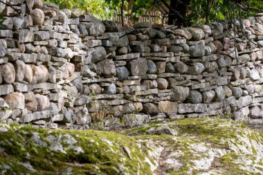 Değişik boyutlarda kayalardan yapılmış taştan bir duvar doğal ve dünyevi bir estetik sergiliyor. Duvar kısmen yosunla kaplı, yeşillik ve ağaçlarla çevrili, sakin bir açık hava ortamı yaratıyor..