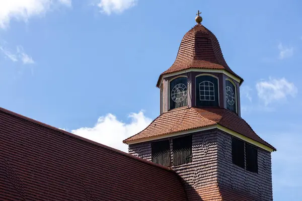 Ayırt edici bir kubbe ve saat yüzlü bir kilise kulesinin yakından görüntüsü, kabarık bulutlu parlak mavi bir gökyüzüne karşı kurulmuş. Yapıda ahşap kiremitler ve tepesinde bir haç var..