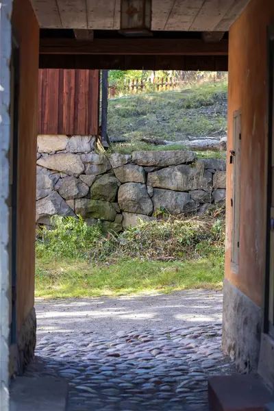 Arka planda taş duvar ve yeşillik olan kaldırım taşlı bir yola açılan bir kemer yolu manzarası. Sahne doğal ışıkla aydınlatılıyor, sakin bir atmosfer yaratıyor..