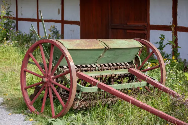 Eski moda bir yeşil ve kırmızı tohum matkabı çimlerin üzerinde dinleniyor, arka planda kırsal ahşap bir bina var. Tohum matkabında büyük ahşap tekerlekler ve tohum ekmek için metal bir mekanizma bulunuyor..