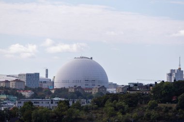 A large white dome structure, known as the Ericsson Globe, surrounded by urban buildings and greenery under a partly cloudy sky. clipart