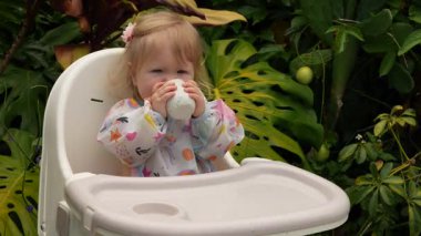 Sevimli beyaz çocuk, açık bir sandalyede açık bir bardaktan içmeyi öğrenirken gülümsüyor. Gerçek bir ebeveyn anı, erken çocukluk gelişimi, samimi bir ifade, doğal bir aile ifadesi.