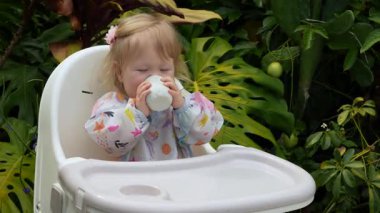 Sevimli çocuk, açık bir bardaktan içmeyi öğrenirken gülümsüyor açık bir sandalyede, gerçek hayatta ebeveynlik anı, erken çocukluk gelişimi, samimi ifade, doğal aile yaşam tarzı