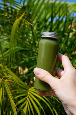 Canlı tropikal yaprakların önünde elinde yeşil detoks suyu şişesi tutuyordu. Sağlıklı yaşam tarzı ve egzotik orman geçmişi üzerine organik beslenme konsepti. Sağlık ve doğal beslenme konsepti.
