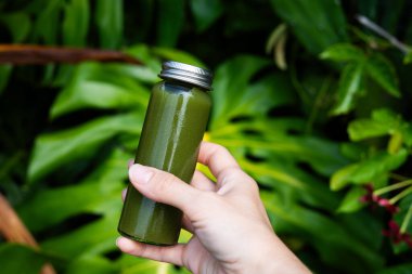Canlı tropikal yaprakların önünde elinde yeşil detoks suyu şişesi tutuyordu. Sağlıklı yaşam tarzı ve egzotik orman geçmişi üzerine organik beslenme konsepti. Sağlık ve doğal beslenme konsepti.