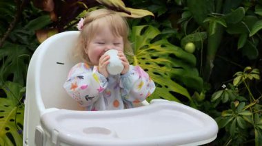 Sevimli çocuk, açık bir bardaktan içmeyi öğrenirken gülümsüyor, açık bir sandalyede, gerçek hayatta ebeveynlik anı, erken çocukluk gelişimi, samimi ifade, doğal aile yaşam tarzı. Yüksek