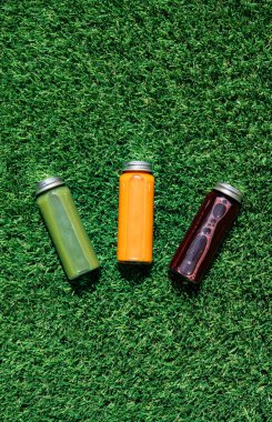 Temiz yemek, sağlıklı yaşam tarzı, fitness pazarlaması, detoks, sağlıklı yaşam için yeşil çimenler üzerinde üç renkli detoks suyu şişesinin dikey resmi.