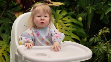 Tatlı çocuk, yemyeşil bir tropikal bahçede plastik bir sandalyeye oturmayı öğreniyor, erken çocukluk dönüm noktası, bebek bağımsızlığı, olağanüstü bir bebek, yemyeşil bir tropik bahçede plastik bir sandalyeye oturmayı öğreniyor.
