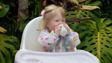 Sevimli çocuk, açık bir bardaktan içmeyi öğrenirken gülümsüyor açık bir sandalyede, gerçek hayatta ebeveynlik anı, erken çocukluk gelişimi, samimi ifade, doğal aile yaşam tarzı