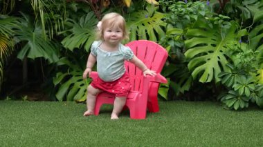 Tatlı çocuk, yemyeşil bir tropikal bahçede plastik bir sandalyeye oturmayı öğreniyor, erken çocukluk dönüm noktası, bebek bağımsızlığı, olağanüstü bir bebek, yemyeşil bir tropik bahçede plastik bir sandalyeye oturmayı öğreniyor.