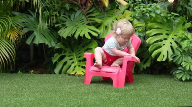 Tatlı çocuk, yemyeşil bir tropikal bahçede plastik bir sandalyeye oturmayı öğreniyor, erken çocukluk dönüm noktası, bebek bağımsızlığı, olağanüstü bir bebek, yemyeşil bir tropik bahçede plastik bir sandalyeye oturmayı öğreniyor.