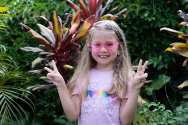 Pembe güneş gözlüğü ve kalp gökkuşağı tişörtlü, uzun sarı saçlı gülümseyen genç kız iki eliyle barış işaretlerini gösteriyor, açık havada renkli yaprakları olan bir tropikal bahçede duruyor. Mutlu çocuk..