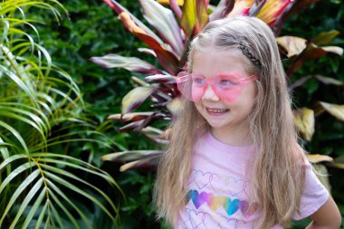 Pembe güneş gözlüklü, uzun sarı saçlı, gülümseyen beyaz bir kız ve açık havada renkli yapraklarla kaplı bir tropik bahçede duran kalp gökkuşağı tişörtü. Mutlu çocuk. Yaz eğlencesi