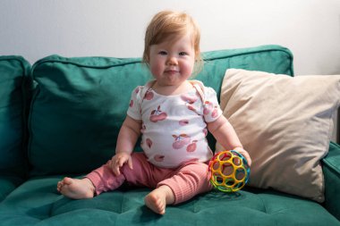 Pembe yanaklı ve açık kahverengi saçlı sevimli bebek bir elinde renkli bir duyusal topla koyu yeşil bir koltukta oturuyor. Bebek rahat ve neşeli bir anın tadını çıkarırken usulca gülümsüyor.. 
