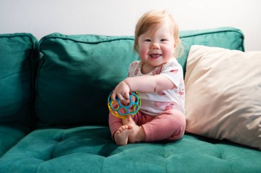 Pembe yanaklı ve açık kahverengi saçlı sevimli bebek bir elinde renkli bir duyusal topla koyu yeşil bir koltukta oturuyor. Bebek rahat ve neşeli bir anın tadını çıkarırken usulca gülümsüyor.. 