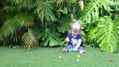 Geniş Canavar Yaprakları ve Palmiye Yapraklarıyla çevrili Lush Tropikal Bahçesinde Yeşil Yapay Çimenler Üzerinde Renkli Oyuncak Bloklarıyla Oynayan Mutlu Kız, Doğal Gün Işığında Gülümseyen ve Dalgalanan