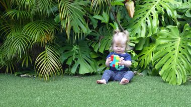 Geniş Canavar Yaprakları ve Palmiye Yapraklarıyla çevrili Lush Tropikal Bahçesinde Yeşil Yapay Çimenler Üzerinde Renkli Oyuncak Bloklarıyla Oynayan Mutlu Kız, Doğal Gün Işığında Gülümseyen ve Dalgalanan