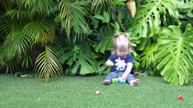 Geniş Canavar Yaprakları ve Palmiye Yapraklarıyla çevrili Lush Tropikal Bahçesinde Yeşil Yapay Çimenler Üzerinde Renkli Oyuncak Bloklarıyla Oynayan Mutlu Kız, Doğal Gün Işığında Gülümseyen ve Dalgalanan