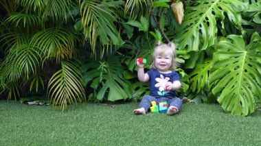 Geniş Canavar Yaprakları ve Palmiye Yapraklarıyla çevrili Lush Tropikal Bahçesinde Yeşil Yapay Çimenler Üzerinde Renkli Oyuncak Bloklarıyla Oynayan Mutlu Kız, Doğal Gün Işığında Gülümseyen ve Dalgalanan