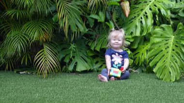 Geniş Canavar Yaprakları ve Palmiye Yapraklarıyla çevrili Lush Tropikal Bahçesinde Yeşil Yapay Çimenler Üzerinde Renkli Oyuncak Bloklarıyla Oynayan Mutlu Kız, Doğal Gün Işığında Gülümseyen ve Dalgalanan