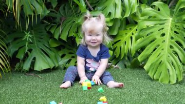Geniş Canavar Yaprakları ve Palmiye Yapraklarıyla çevrili Lush Tropikal Bahçesinde Yeşil Yapay Çimenler Üzerinde Renkli Oyuncak Bloklarıyla Oynayan Mutlu Kız, Doğal Gün Işığında Gülümseyen ve Dalgalanan