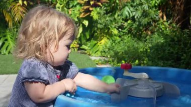 Mavi bir açık hava su masasında oyuncak ve yüzen nesnelerle oynayan meraklı bir kız çocuğu, yemyeşil bir bahçede, su oyunlarını ve dokunma, hareket ve konsantrasyon yoluyla duyusal gelişimi keşfediyor.