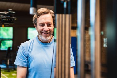 Bir adam gülümsüyor ve spor salonunda bir ağırlık makinesinin önünde duruyor. Trisepslerini çalıştırıyor. Mavi bir gömlek giyiyor ve mutlu.