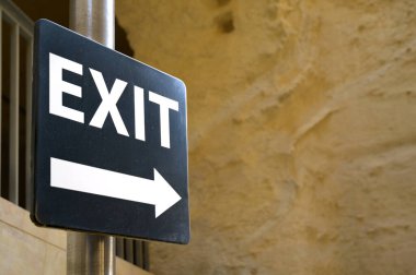 Çıkış işaretinde ok sağa, siyah zemin üzerinde beyaz harfler arka planda taş duvar, Valetta, Malta.