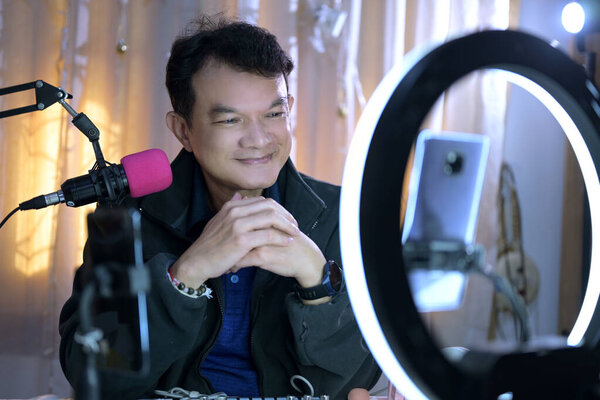 Мужчина среднего возраста живет в стремлении через социальные сети, такие как Tiktok или Facebook. Он использует смартфон, соединенный с круговой светодиодной лампой, с микрофоном и звуковой картой..