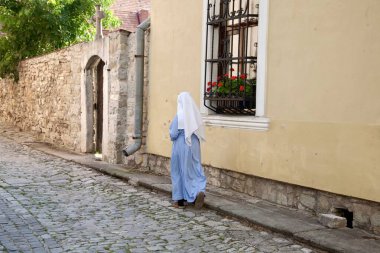 Fotoğraf, beyaz ve mavi elbiseli bir rahibenin eski bir bina boyunca yürüdüğünü gösteriyor. Onun sakin ve sakin yürüyüşü bir barış ortamı yaratıyor..