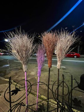 Akşamları yolun yukarısındaki çitin altında dört farklı renkte süpürge duruyor. Arka planda arabalar ve evler görülüyor..