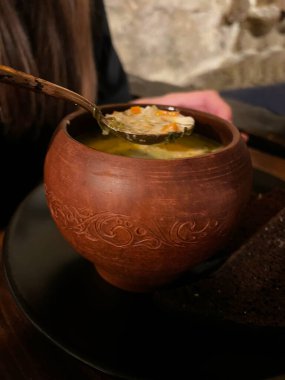 Fotoğrafta taze ısırgan otlarıyla süslenmiş zengin bir sebze çorbası var. Yakınlarda büyük bir kaşık uzanıyor, ve pembe şarap kadehleri ahşap bir masada duruyor, rahat bir atmosfer yaratıyor..