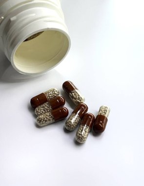 Brown pharmaceutical capsules with a transparent shell containing microgranules lie on a white surface next to an open plastic jar. clipart